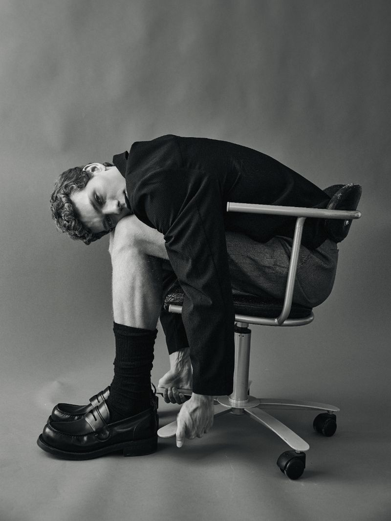 Model Caesar van den Idsert hunched over on a swivel office chair wearing a dark jacket and loafers against a grey studio backdrop.
