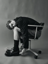 Model Caesar van den Idsert hunched over on a swivel office chair wearing a dark jacket and loafers against a grey studio backdrop.