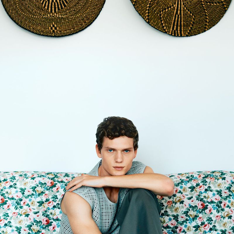 Model Caesar van den Idsert seated in a sleeveless mesh knit top against a floral sofa and white wall with woven decor.