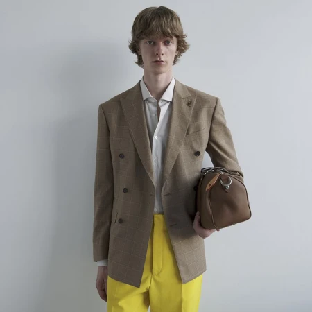 Model Guus Bracke in a tan plaid double-breasted blazer and bright yellow trousers holding a brown leather bag against a neutral backdrop.