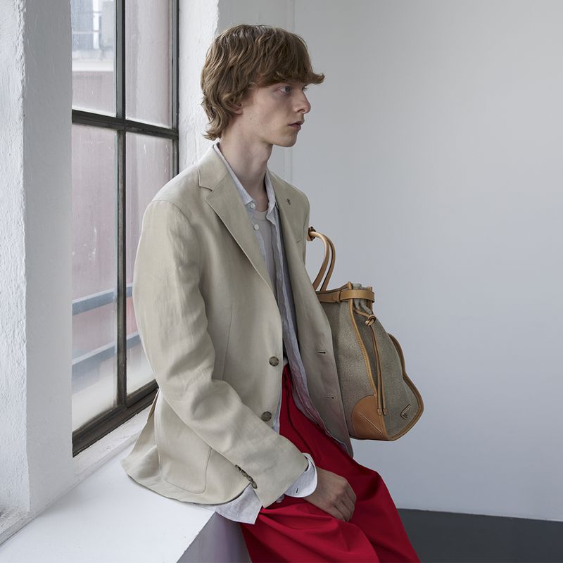 Model Guus Bracke seated on a white windowsill wearing a beige linen blazer and bright red trousers next to a canvas tote bag.