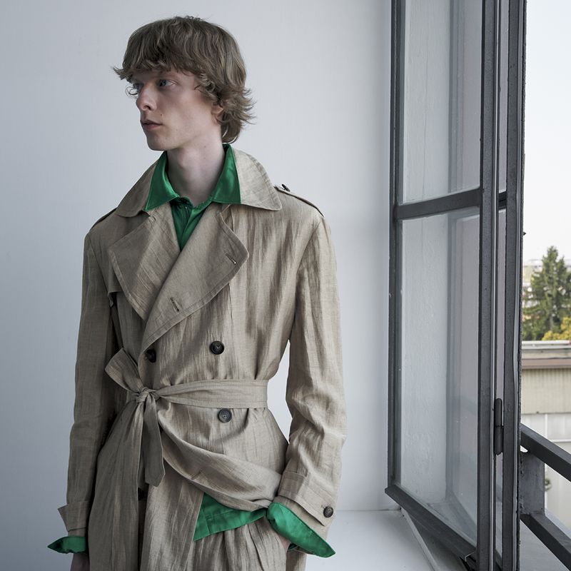Model Guus Bracke wearing a beige linen belted trench coat over a vivid green collared shirt beside a tall window.