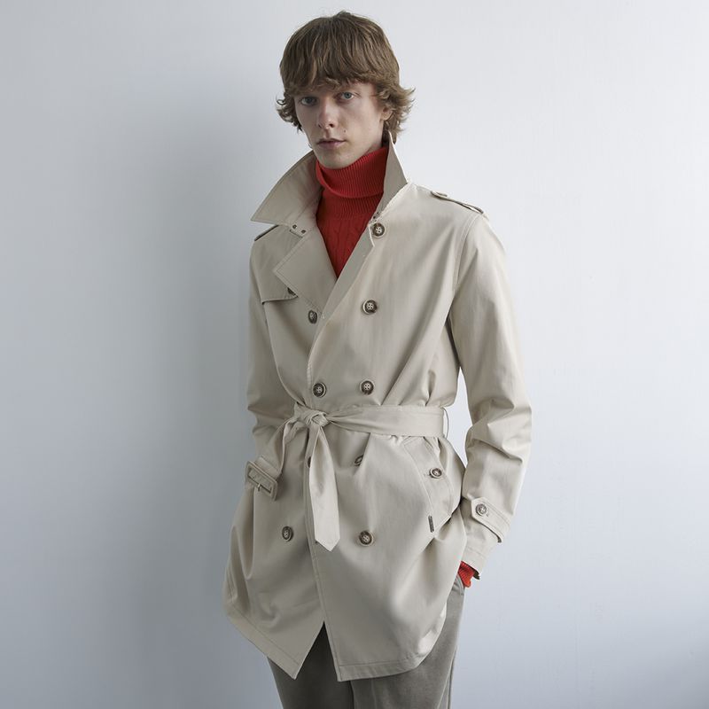 Model Guus Bracke posing in a belted beige trench coat and red turtleneck sweater against a plain white studio backdrop.