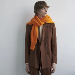 Model Guus Bracke wearing a brown double-breasted suit and a bright orange knit scarf against a clean studio backdrop.