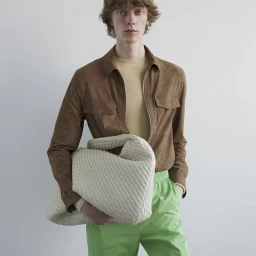 Model Guus Bracke holding a large woven cream bag wearing a brown suede shirt and lime green pants against a white background.