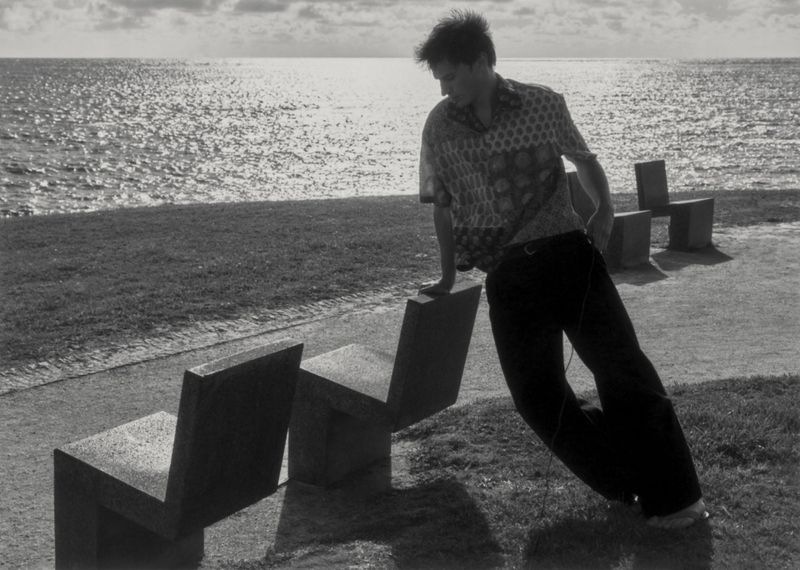 Man in a printed short-sleeve shirt and dark trousers leaning on a concrete park chair overlooking the ocean in black and white.