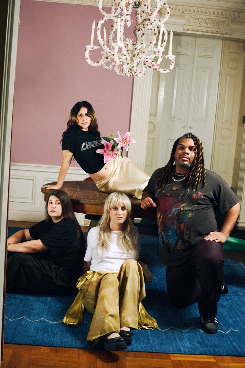 Four people posing together in an interior room with a decorated chandelier, pink wall, and blue carpet for W Magazine.