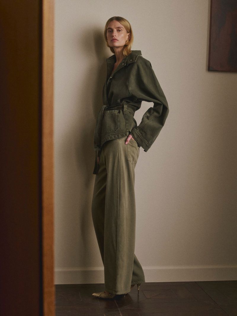 Model in olive green belted jacket and matching trousers posing against a neutral wall for DL1961 Pre-Fall 2026.