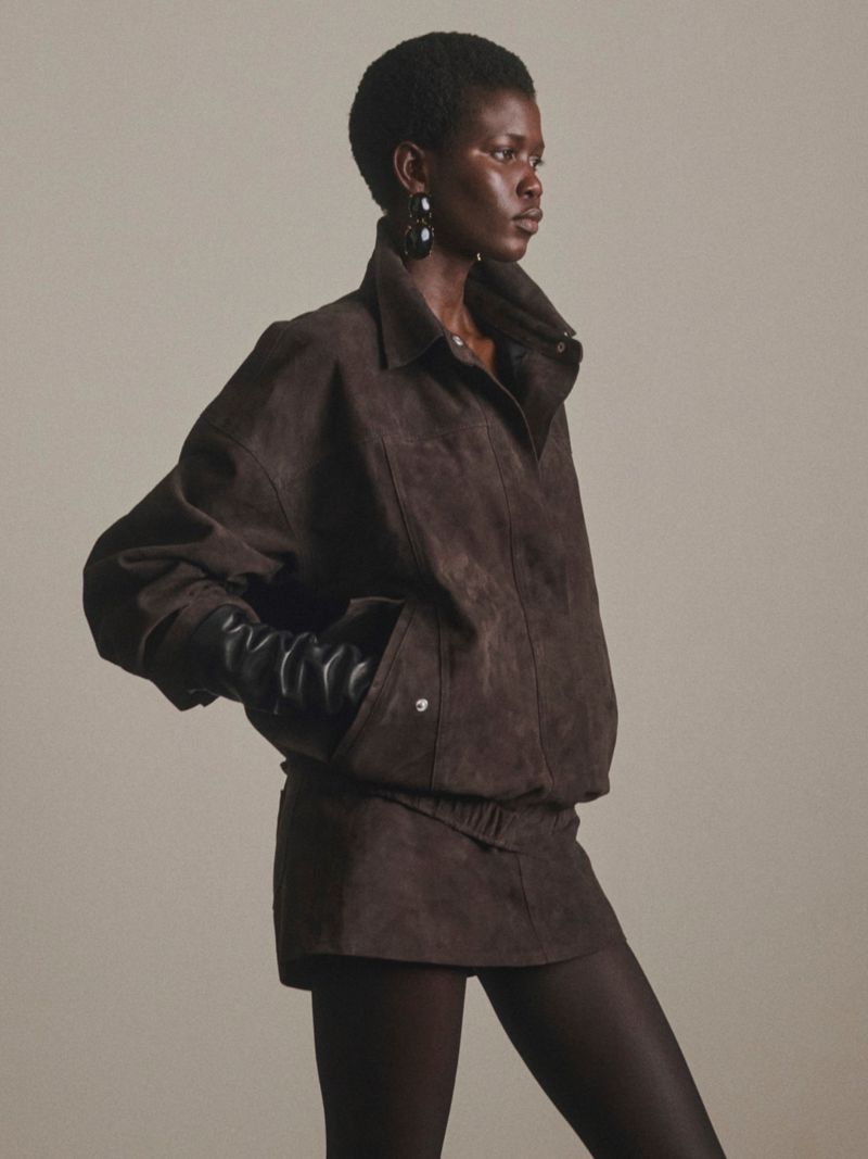 Model Ajah Angau Jok in a dark brown suede jacket and matching skirt with black leather gloves against a studio backdrop.