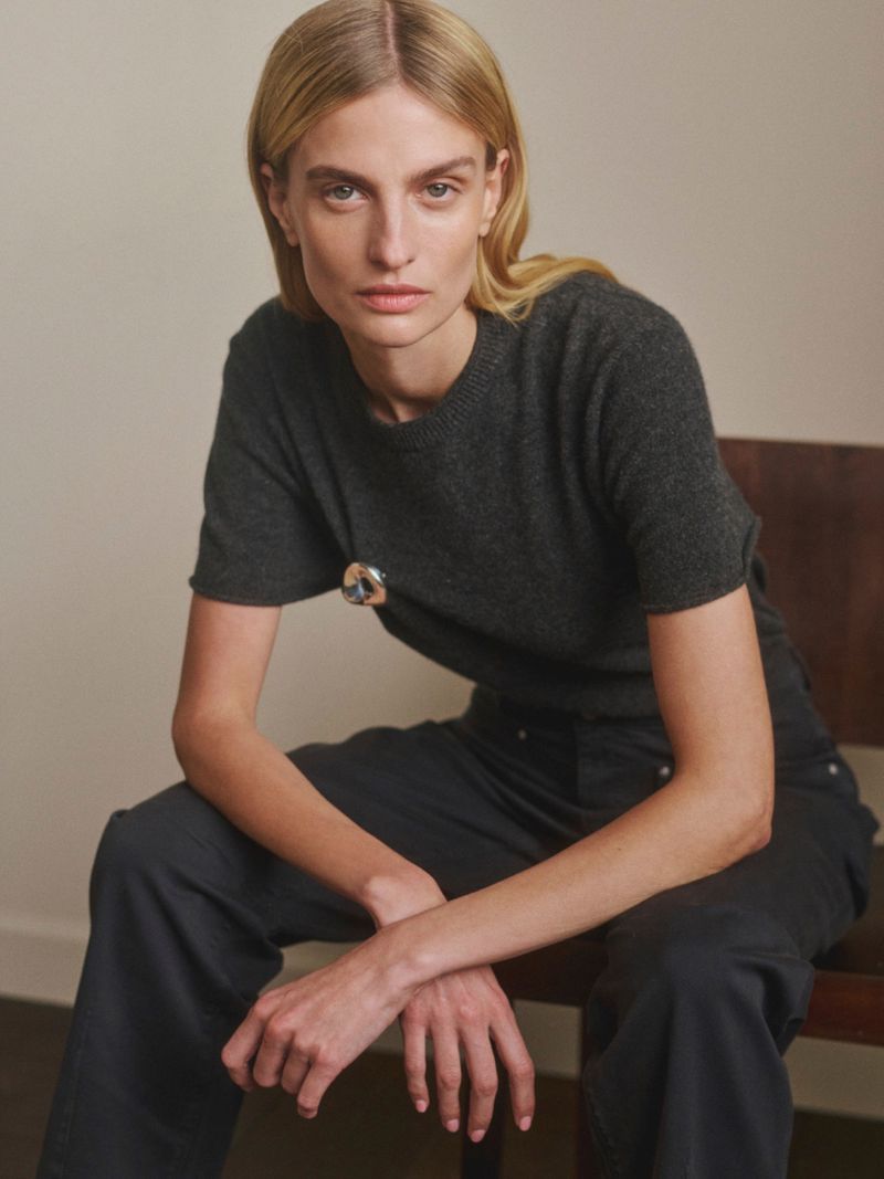 Veronika Vilim seated on a wooden chair wearing a charcoal knit top and dark pants for DL1961 Pre-Fall 2026.