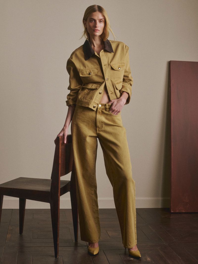 Veronika Vilim in a mustard denim jacket and wide-leg trousers standing beside a dark wooden chair against a neutral studio wall.