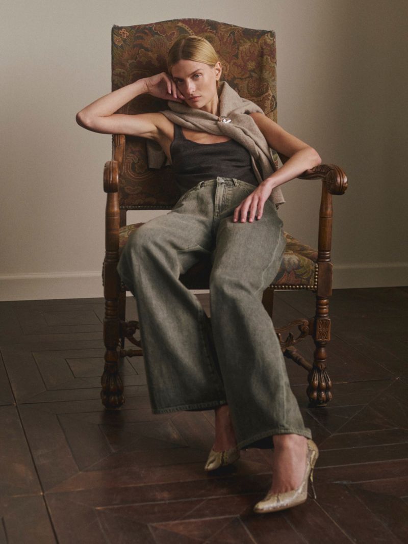 Veronika Vilim seated in an upholstered vintage armchair wearing wide-leg denim jeans, a black tank top, and a beige knit sweater.
