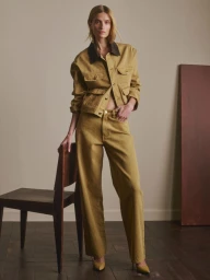 Veronika Vilim in a mustard denim jacket and wide-leg trousers standing beside a dark wooden chair against a neutral studio wall.