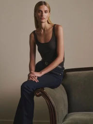 Veronika Vilim sitting on the arm of a green velvet sofa wearing a dark scoop-neck tank top and dark blue denim flare jeans.