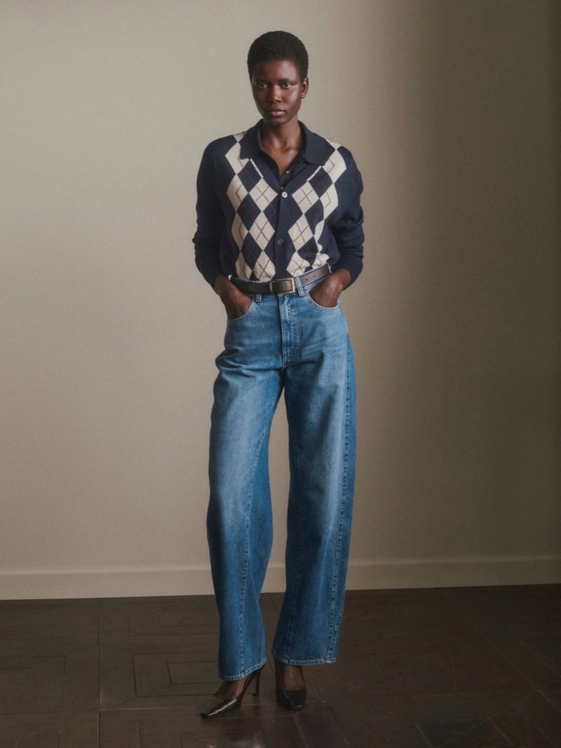 Model Ajah Angau Jok standing in an argyle knit cardigan, high-waisted wide-leg blue jeans, and dark pointed heels.