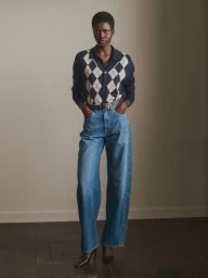 Model Ajah Angau Jok standing in an argyle knit cardigan, high-waisted wide-leg blue jeans, and dark pointed heels.