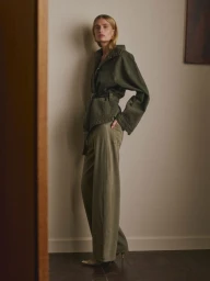 Model in olive green belted jacket and matching trousers posing against a neutral wall for DL1961 Pre-Fall 2026.