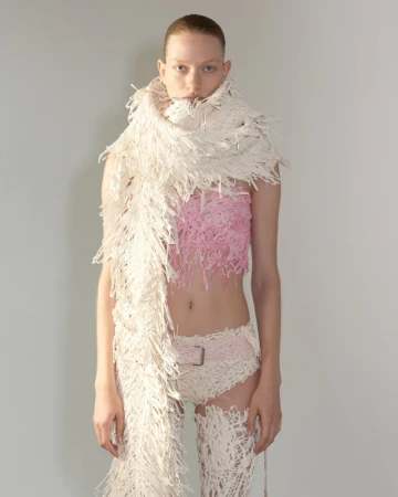 Model wearing a textured cream fringed scarf, pink fringed top, and white briefs against a light grey studio background.