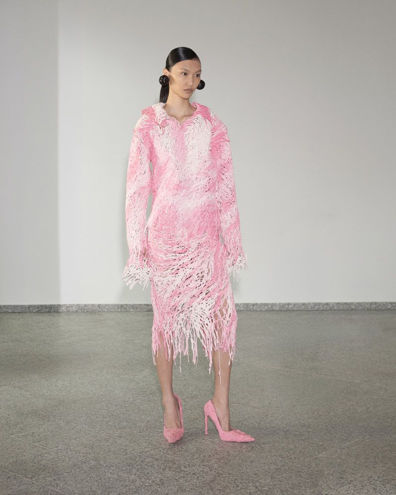 Model wearing a textured pink and white fringed long-sleeve dress and matching pink pointed-toe stiletto heels in a studio.