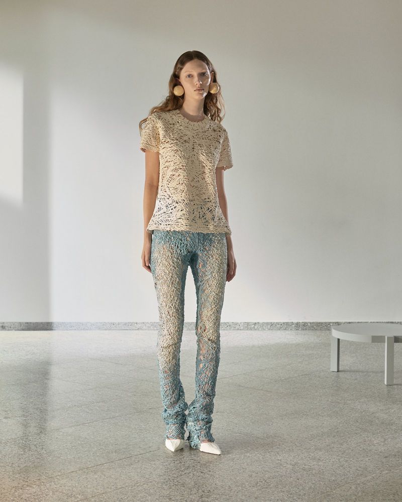 Model wearing a cream textured top and light blue lace pants with large spherical earrings in a minimalist interior setting.