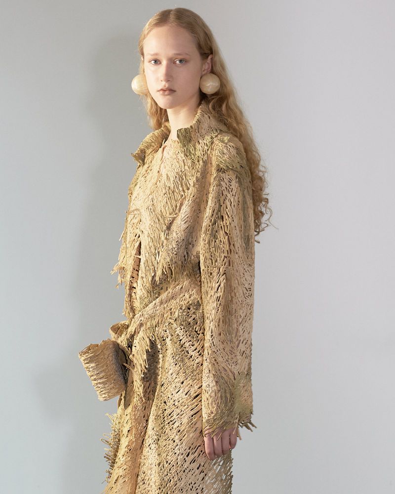 Model in a textured beige frayed coat and large spherical earrings posing against a plain grey studio backdrop.
