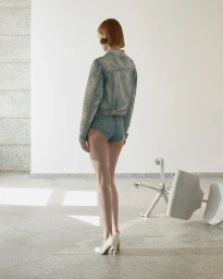 A model seen from behind wearing a mottled blue jacket and matching shorts with white high heels near an overturned office chair.