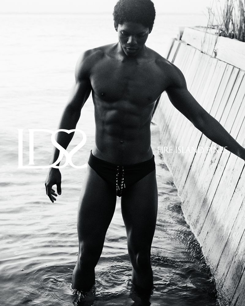 Monochrome portrait of a muscular Black male model in a swimsuit, standing in water next to a wooden structure, creating a striking visual.