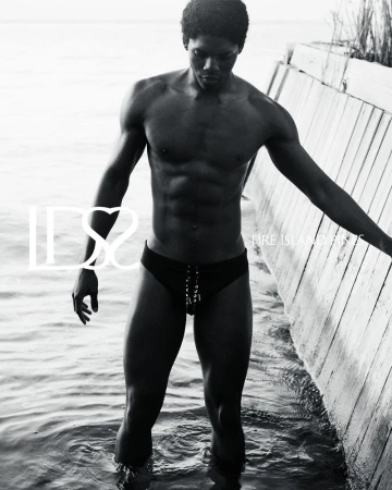 Monochrome portrait of a muscular Black male model in a swimsuit, standing in water next to a wooden structure, creating a striking visual.