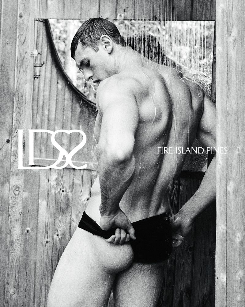Monochrome image of a muscular man under an outdoor shower with water running, wearing swim trunks, standing against a rustic wooden wall.