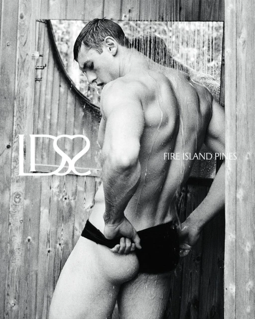 Monochrome image of a muscular man under an outdoor shower with water running, wearing swim trunks, standing against a rustic wooden wall.