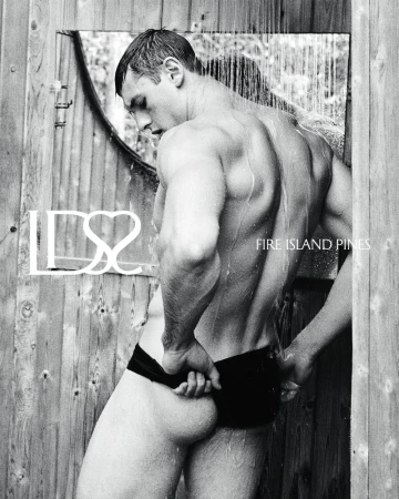 Monochrome image of a muscular man under an outdoor shower with water running, wearing swim trunks, standing against a rustic wooden wall.