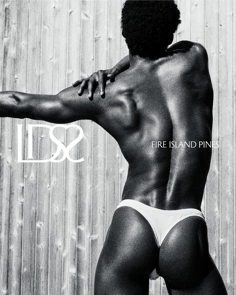 Monochrome portrait of a muscular adult male posing in a white swimsuit against a wood wall backdrop, evoking a fashion editorial.