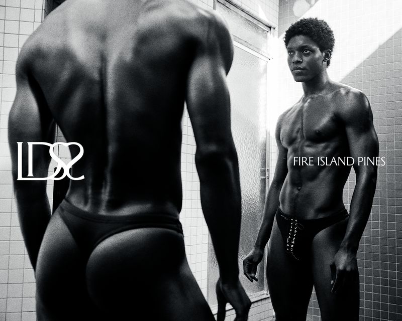 Monochrome photo of muscular Black man in swimsuit standing in front of bathroom mirror in underwear, showcasing fitness and physique.