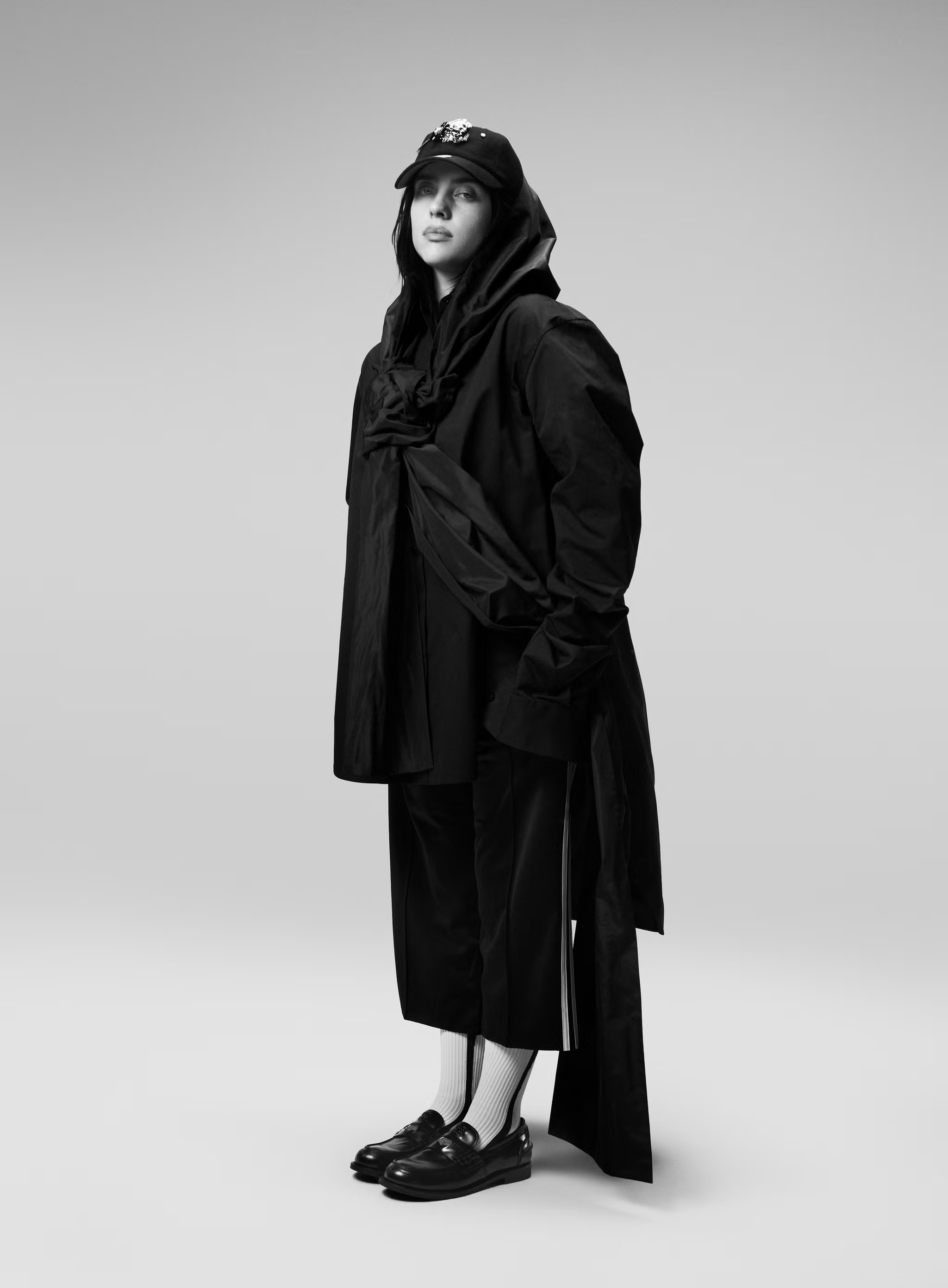 Billie Eilish standing in an oversized black draped hooded coat, cap, cropped trousers, white socks, and loafers against a studio backdrop.