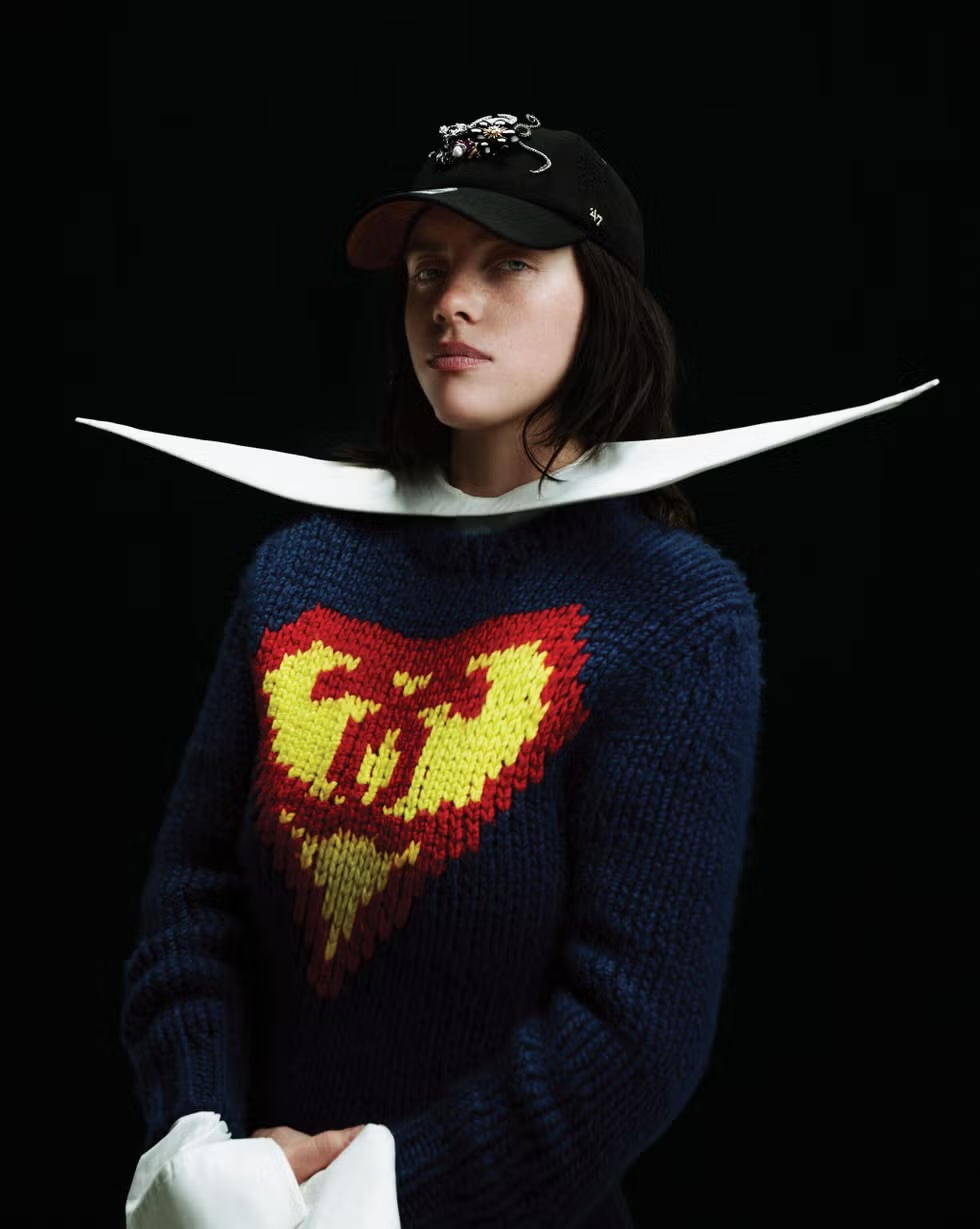 Billie Eilish wearing an embellished baseball cap, wide sculptural white collar, and navy graphic knit sweater on a black backdrop.