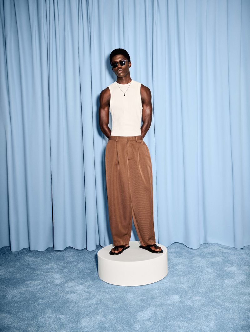 Black man wearing sunglasses, a white sleeveless tank top, and wide brown trousers standing on a round plinth in front of a blue curtain.