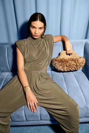 Woman seated on a light blue channel-tufted sofa wearing an olive green sleeveless jumpsuit and holding a fringed straw clutch.