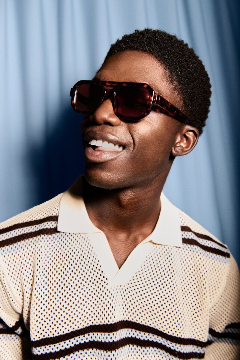 Black man smiling in tortoiseshell sunglasses and a cream knit striped polo shirt against a blue curtain backdrop.