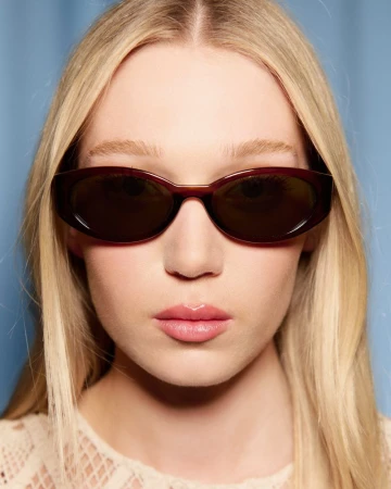 Close-up portrait of a blonde woman wearing dark oval sunglasses and an off-white crochet top against a blue background.