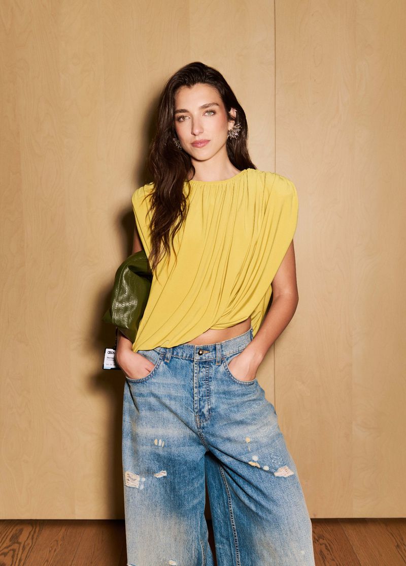 Model in draped yellow top and distressed wide-leg jeans holding an olive green bag against a wood panel wall.
