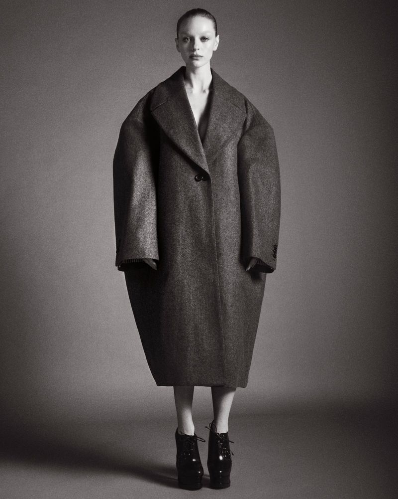 Olivia Rose Keegan posing in an oversized structured grey cocoon coat and black platform boots against a seamless grey background.