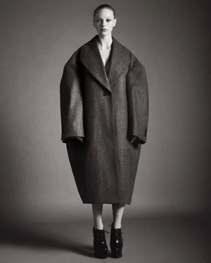 Olivia Rose Keegan posing in an oversized structured grey cocoon coat and black platform boots against a seamless grey background.