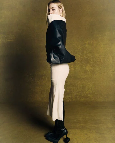 Olivia Rose Keegan posing in profile wearing a black leather jacket, cream slit skirt, and sculptural heels against an olive backdrop.