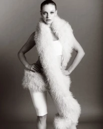 Olivia Rose Keegan posing with hands on hips wearing a voluminous white textured boa, crop top, and shorts in studio.