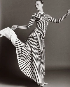 Olivia Rose Keegan poses dynamically wearing an optical illusion striped maxi dress and white boots against a grey studio background.