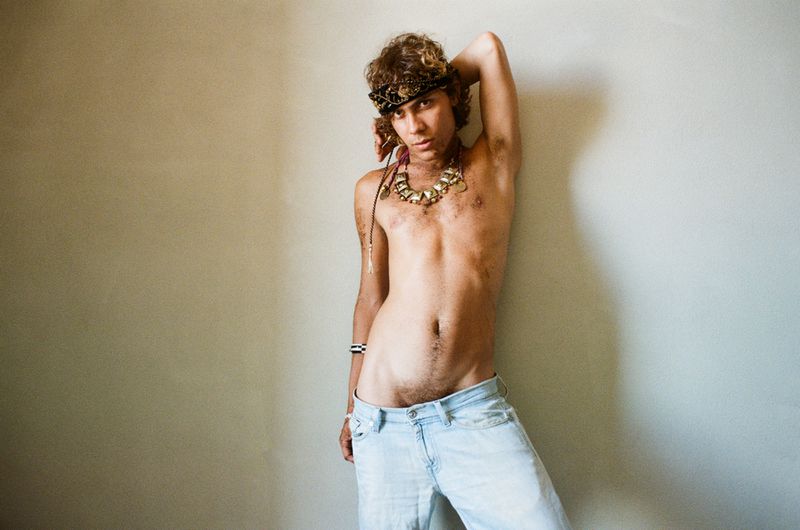 Shirtless person wearing a patterned headband, chunky necklace, and light wash denim jeans leaning against a wall.