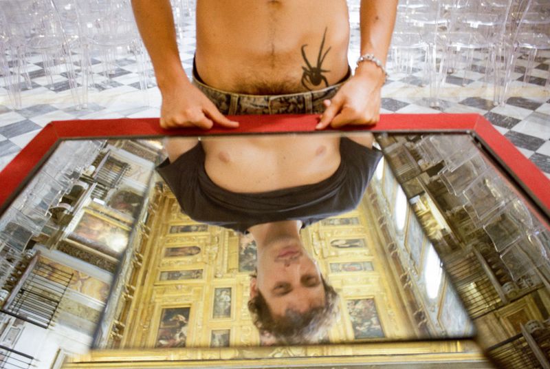 Man holding a red-framed mirror showing his reflection against an ornate gallery ceiling, with a spider tattoo visible on his torso.