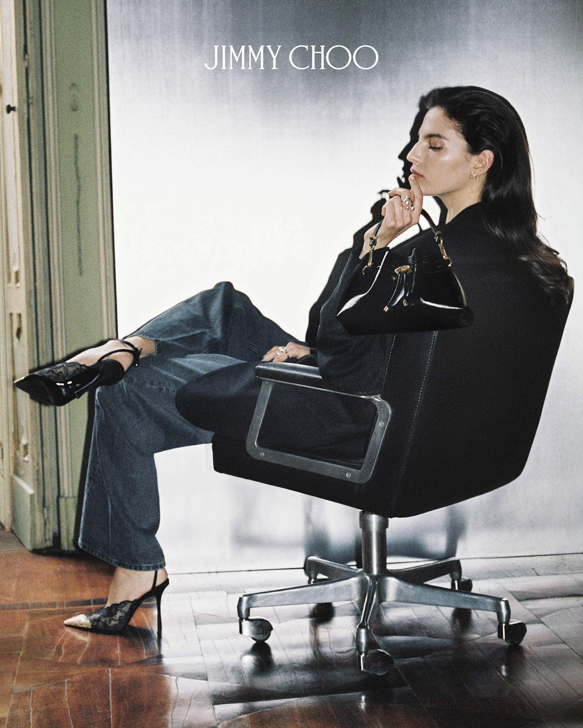 A woman seated in a black office chair holding a handbag, wearing a dark blazer, denim jeans, and black slingback heels.