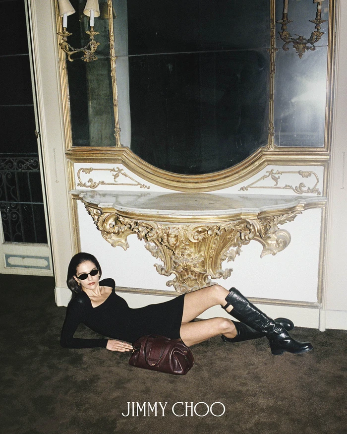 Model reclining on carpet in black mini dress, knee-high boots, and sunglasses with a burgundy handbag in front of a gilded console.