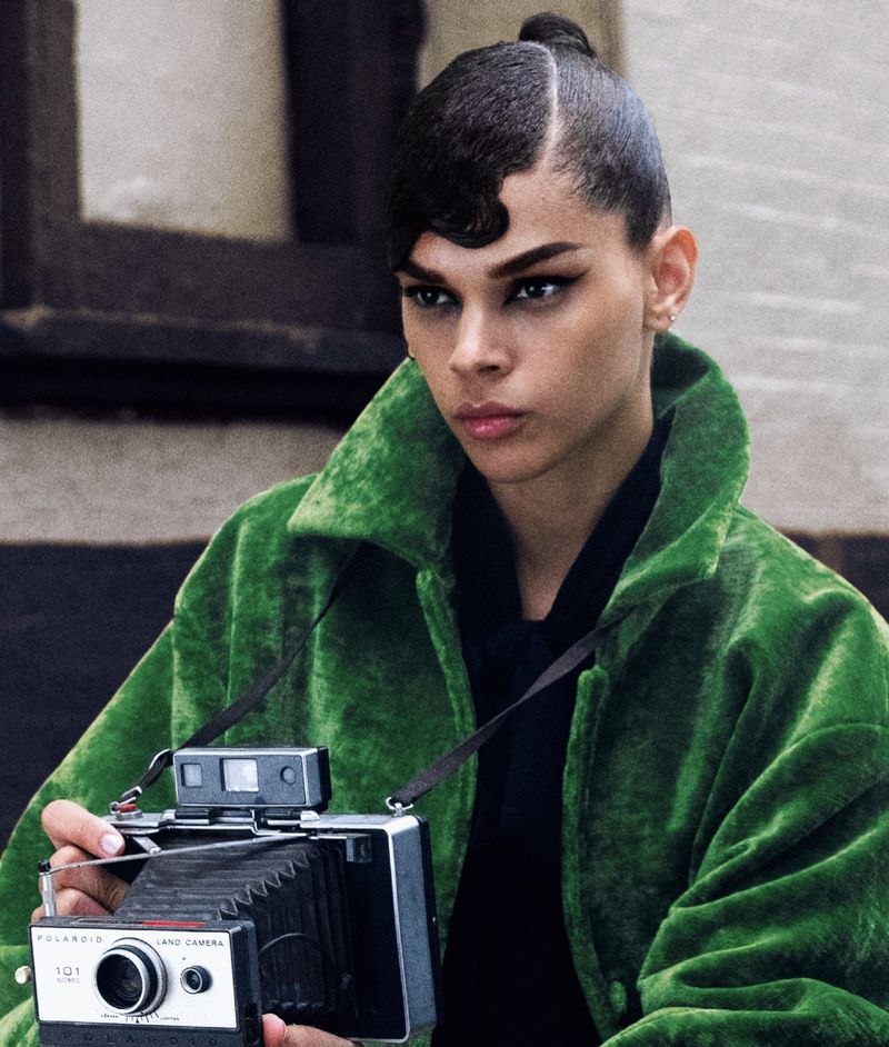 Woman in green jacket poses with vintage Polaroid camera in outdoor setting with light grey background, creative hair and dark makeup.